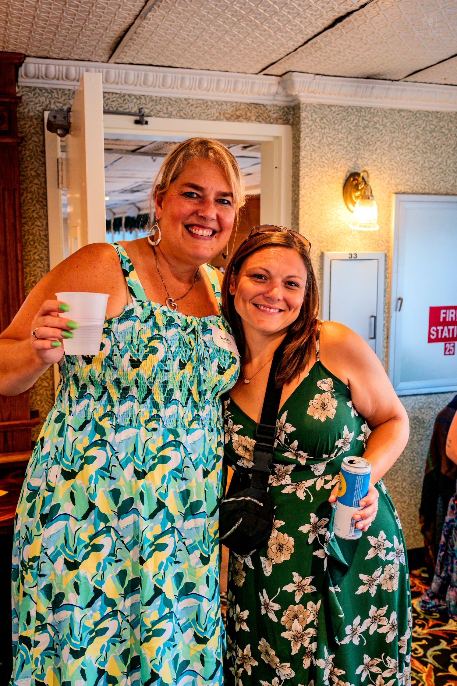 Two smiling women in floral dresses holding drinks pose together at an indoor social event.