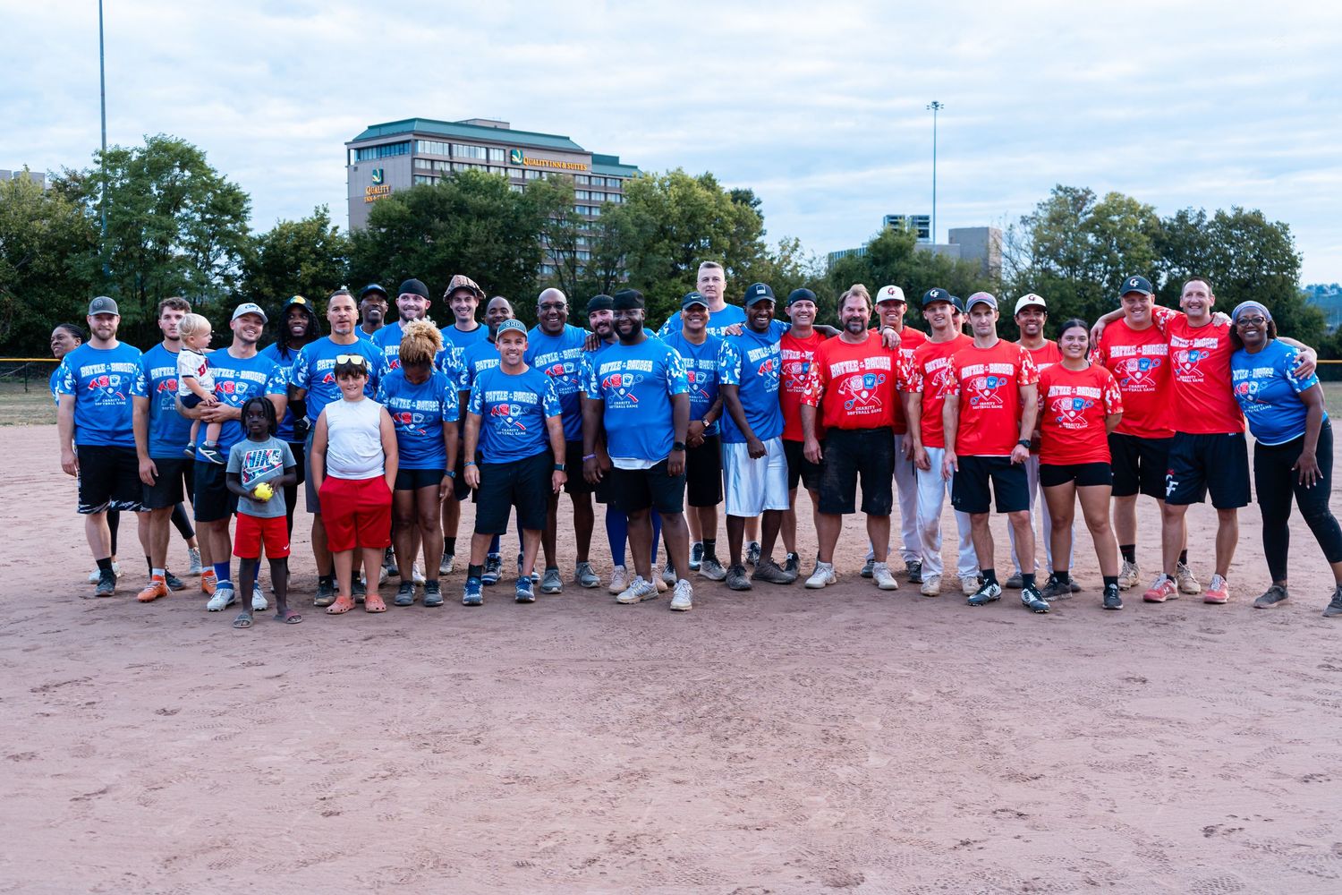 Two teams in blue and red jerseys pose together on a softball field for a group photo after a game.