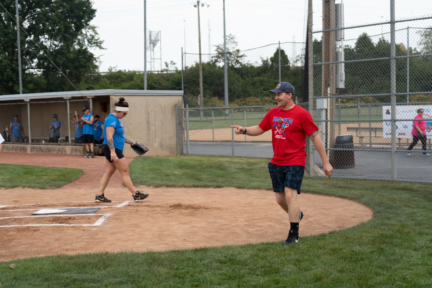 Players compete in a recreational softball game on a dirt infield diamond with green fencing and trees in background.