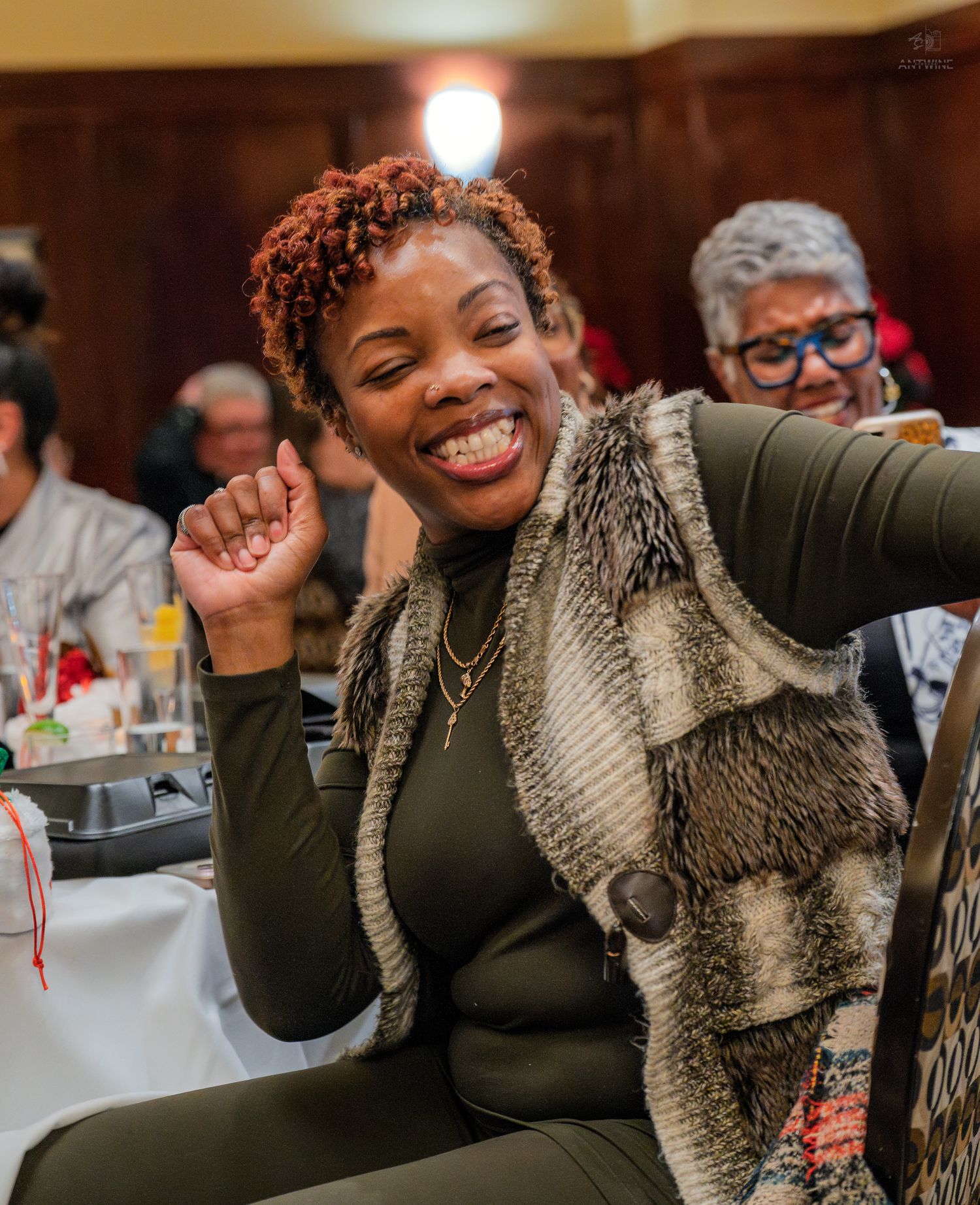 Joyful woman in fur vest smiling and gesturing at a social gathering.