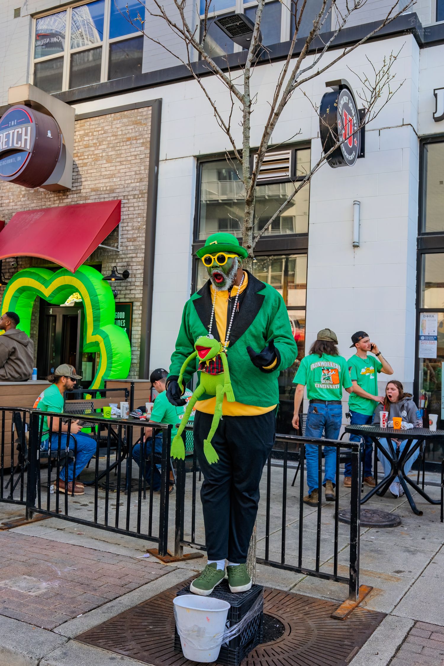 Man in green St. Patrick's Day outfit holding Kermit the Frog doll stands on bucket outside festive bar.