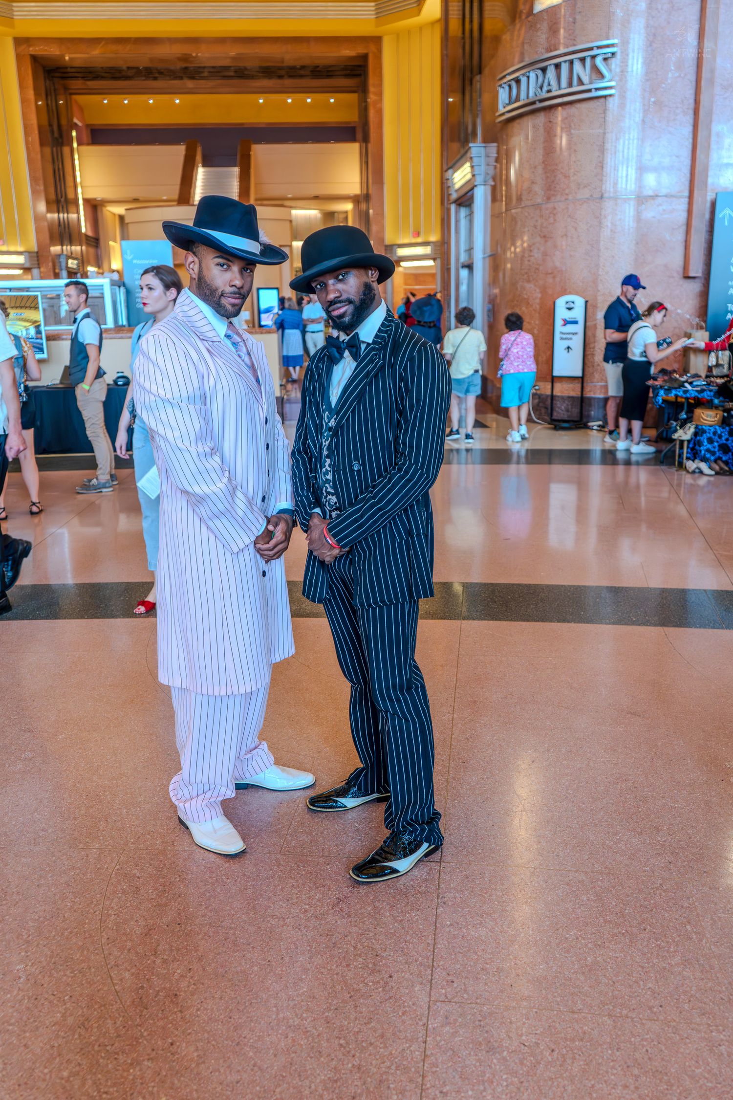 Two men in pinstripe zoot suits and fedora hats pose inside a grand train station.