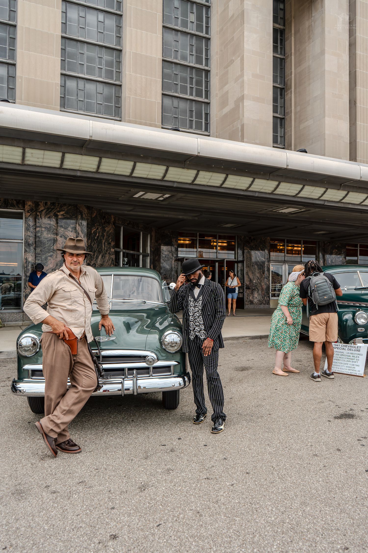 Two retro-dressed men lean on a classic green vintage car outside a large historic building.