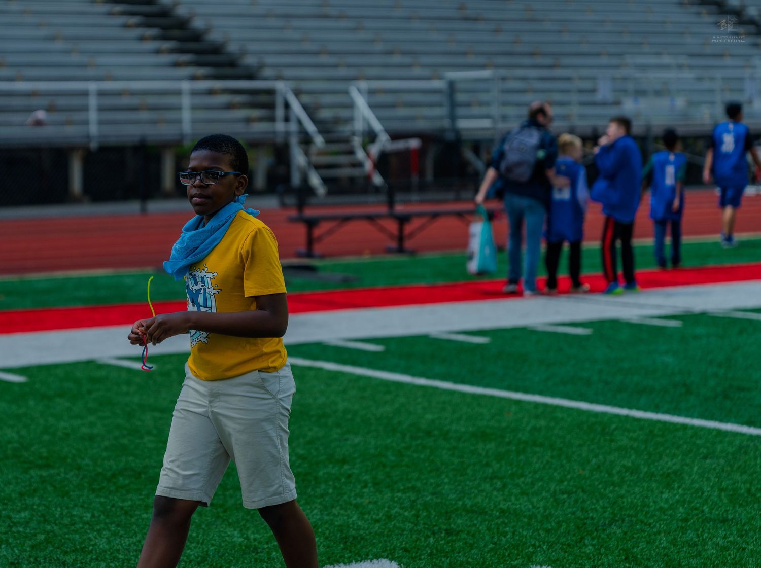 A young person wearing a yellow shirt and blue scarf stands on a red track field at a stadium.