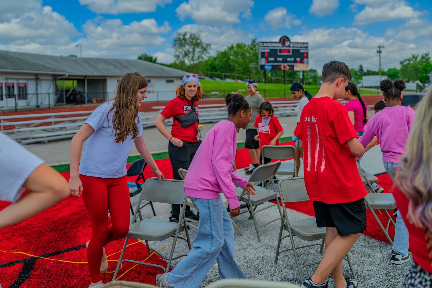 Students in red and pink shirts set up folding chairs outdoors near a scoreboard on a sunny day.