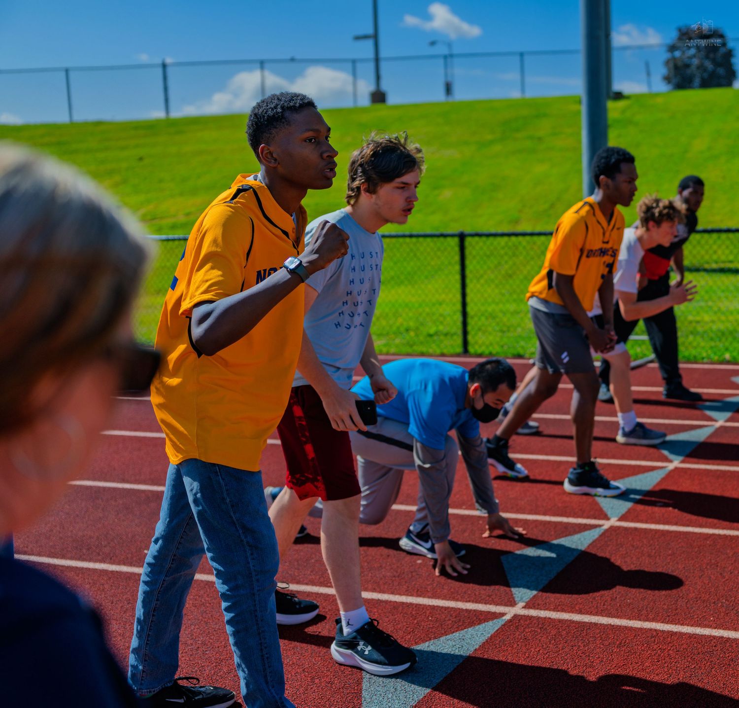 Students in yellow shirts line up at starting blocks on a red running track during an outdoor track and field event.