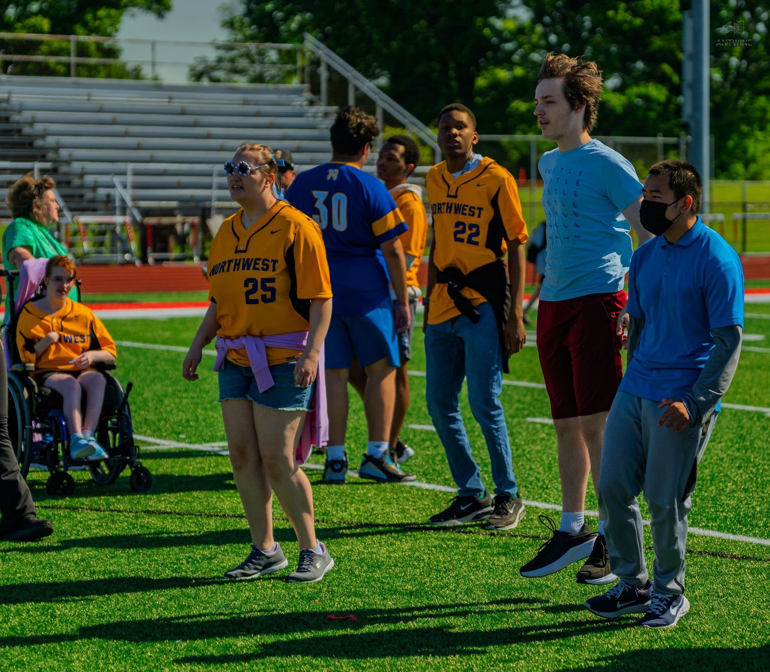 Students in yellow jerseys with numbers 25 and 22 stand on a green sports field during an outdoor school athletic event.