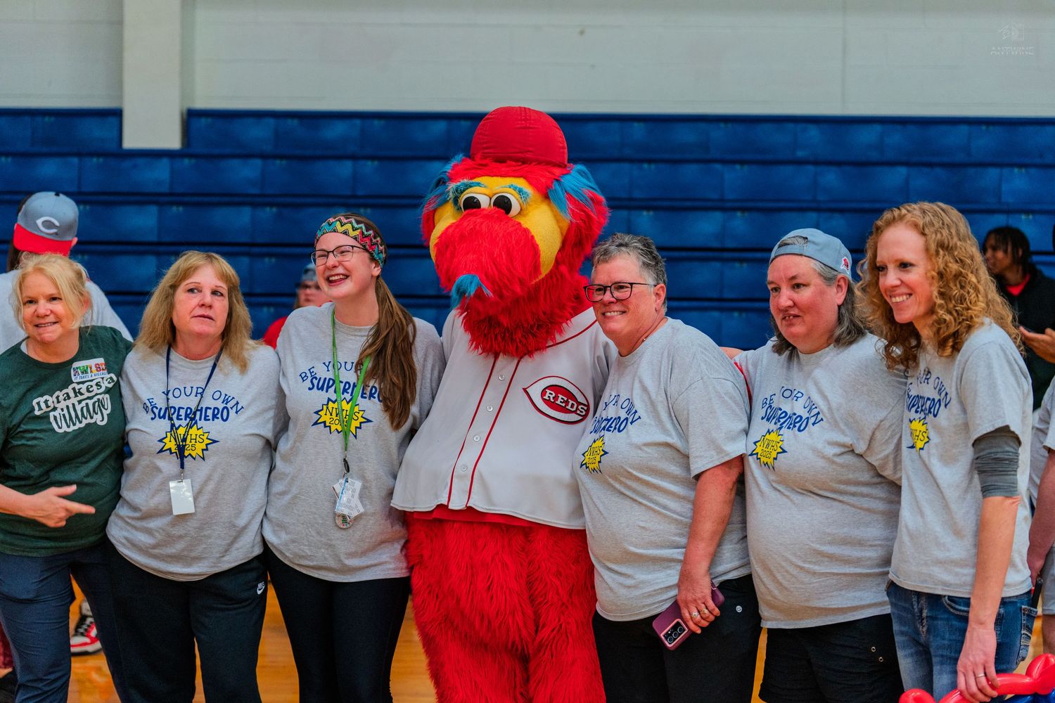Group of women posing with Cincinnati Reds mascot Mr. Red in a gymnasium, wearing matching gray t-shirts.