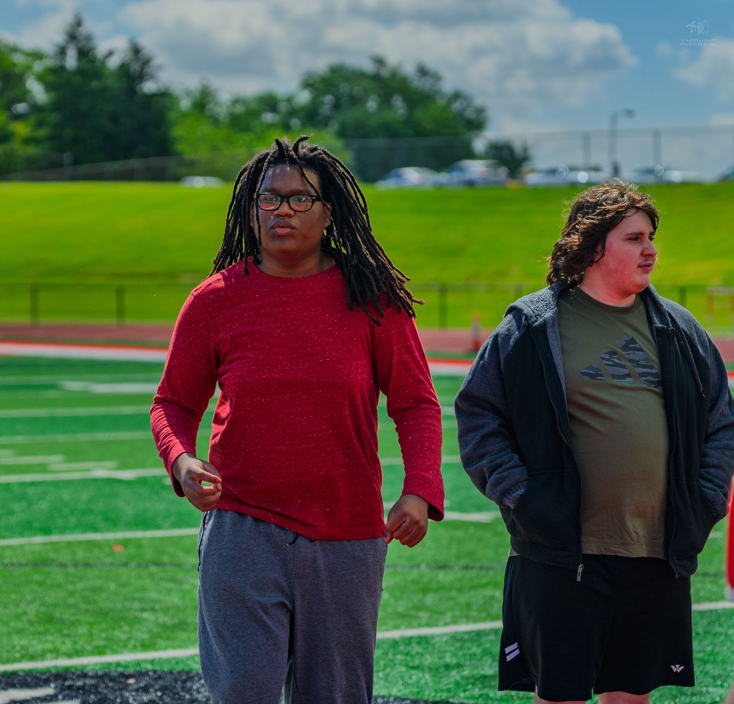 Two athletes stand on a green turf track field, one in a red long-sleeve shirt and another in a dark jacket.
