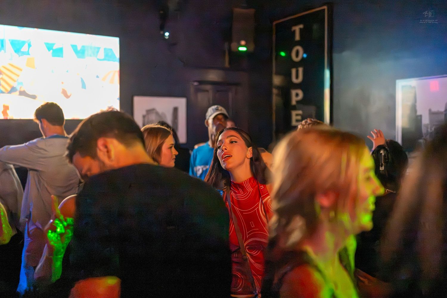 Young adults dancing in a dimly lit nightclub with colorful lights and a projection screen.
