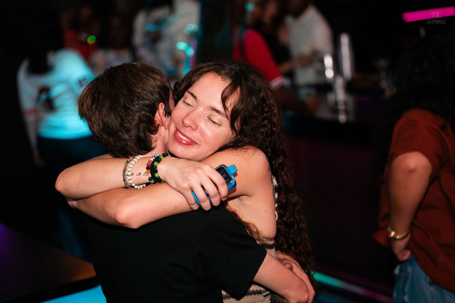 A smiling curly-haired woman joyfully hugs someone under blue lights on a nightclub dance floor.