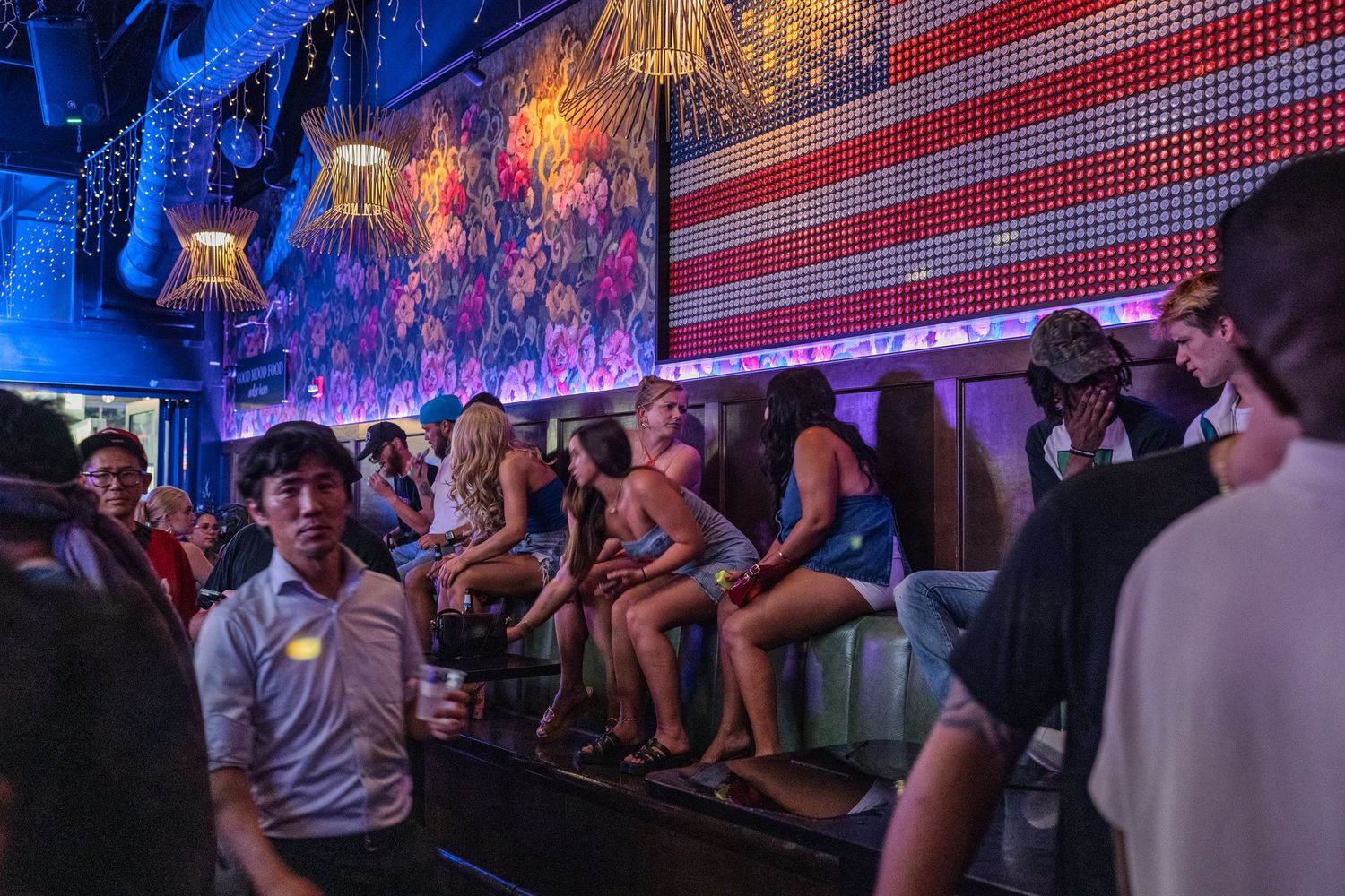 Lively nightclub scene with American flag mural, blue neon lights, and crowd of partygoers.