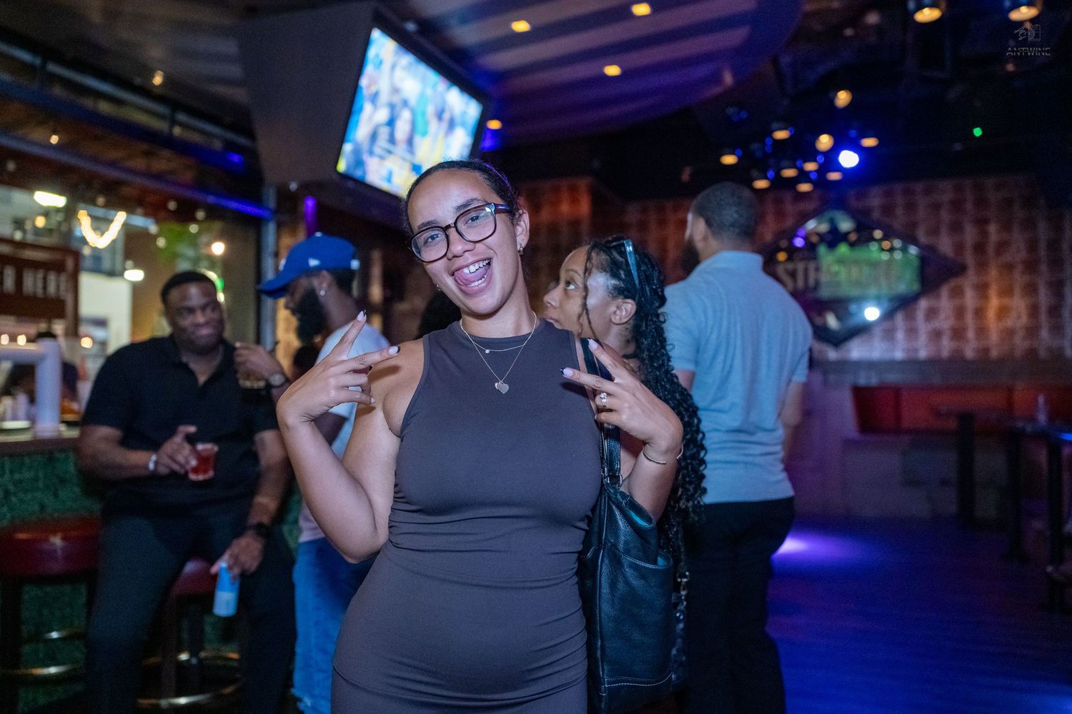 Smiling woman with glasses poses with peace signs in a crowded nightclub bar environment.