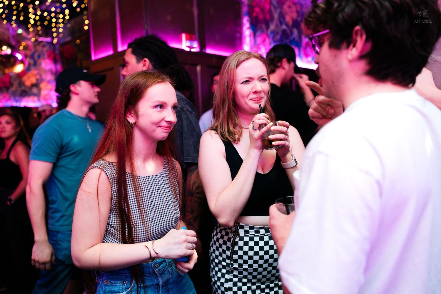 Two young women smiling and holding drinks at a crowded nightclub event.