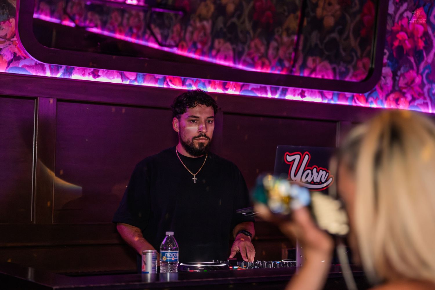 DJ performing at a nightclub booth under purple neon lights and floral decor.