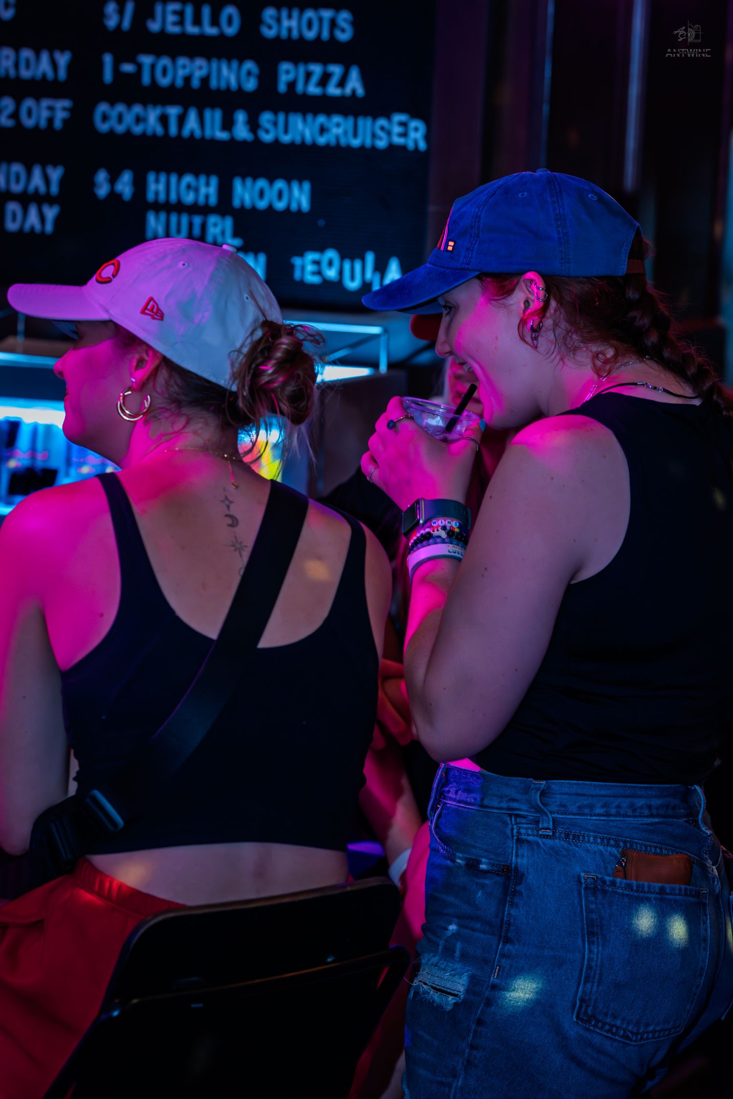 Two women in baseball caps drinking and chatting at a neon-lit bar at night.