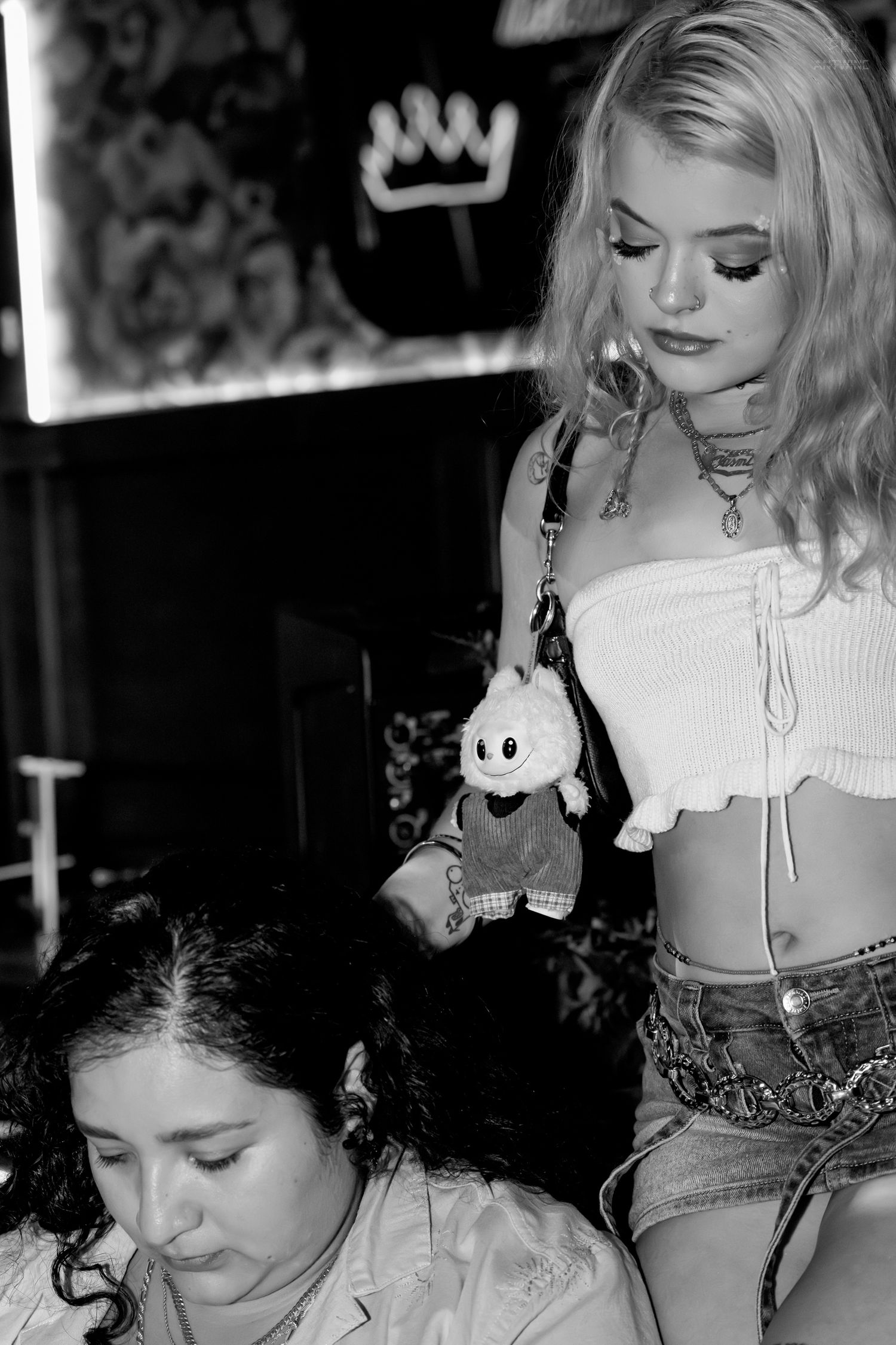 Black and white photo of two women in nightclub, one standing with stuffed toy bag charm.