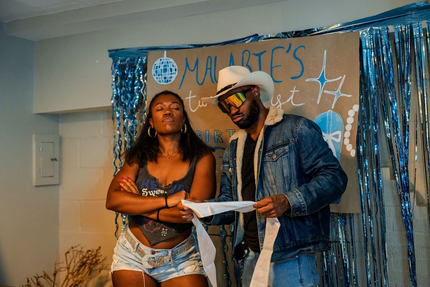 Couple looking at a birthday sash together in front of a blue party backdrop.