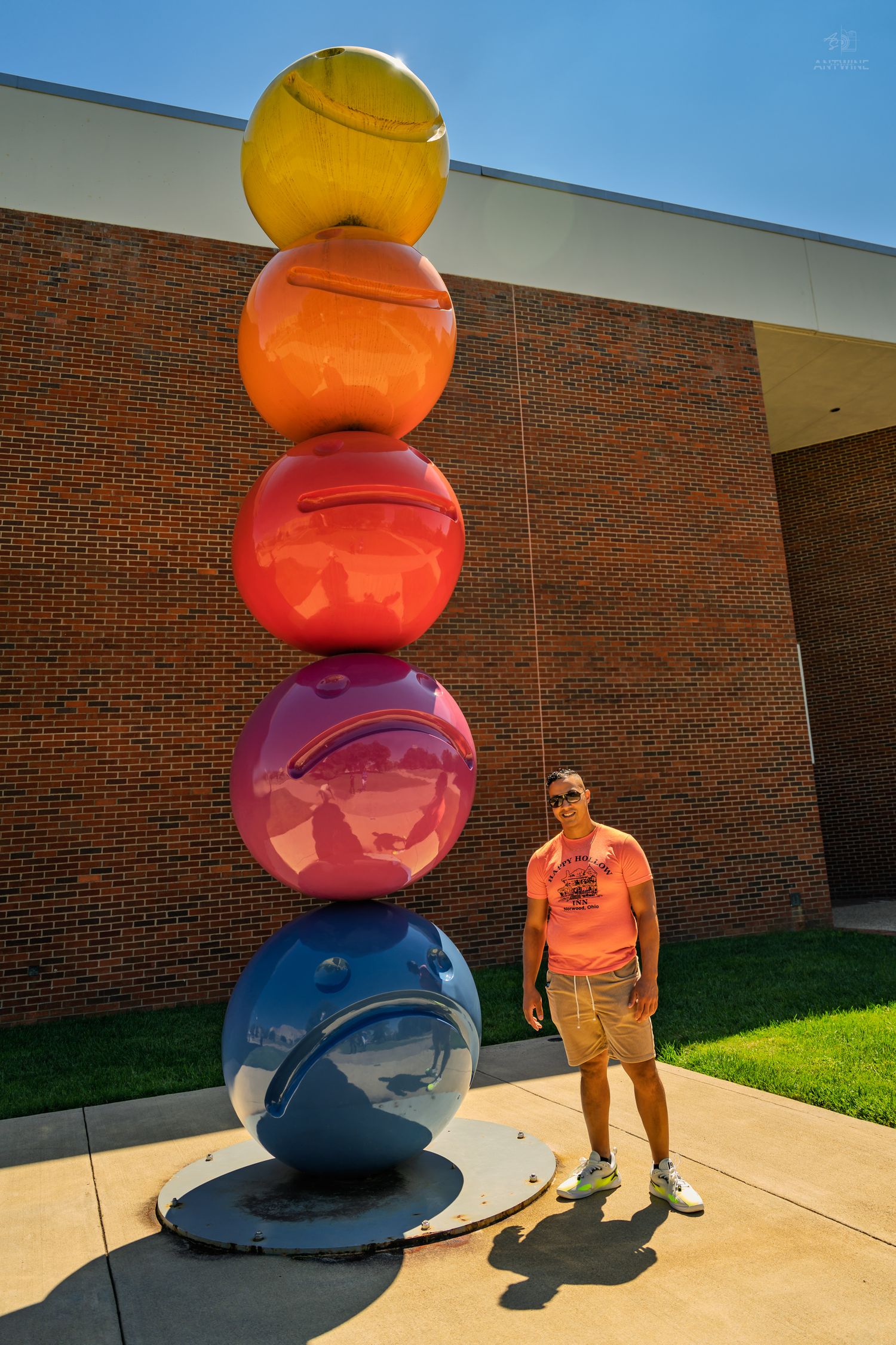 Man in pink shirt standing beside tall rainbow-colored stacked spheres sculpture outside brick building.