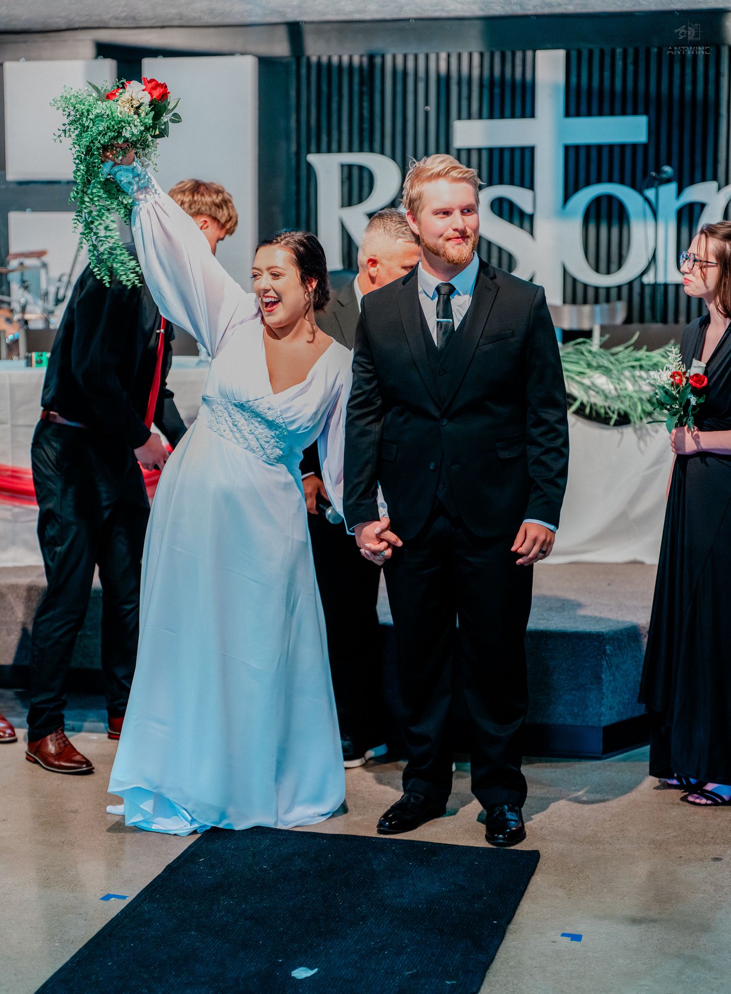 Excited bride raises bouquet while holding grooms hand after wedding ceremony in front of cross backdrop.