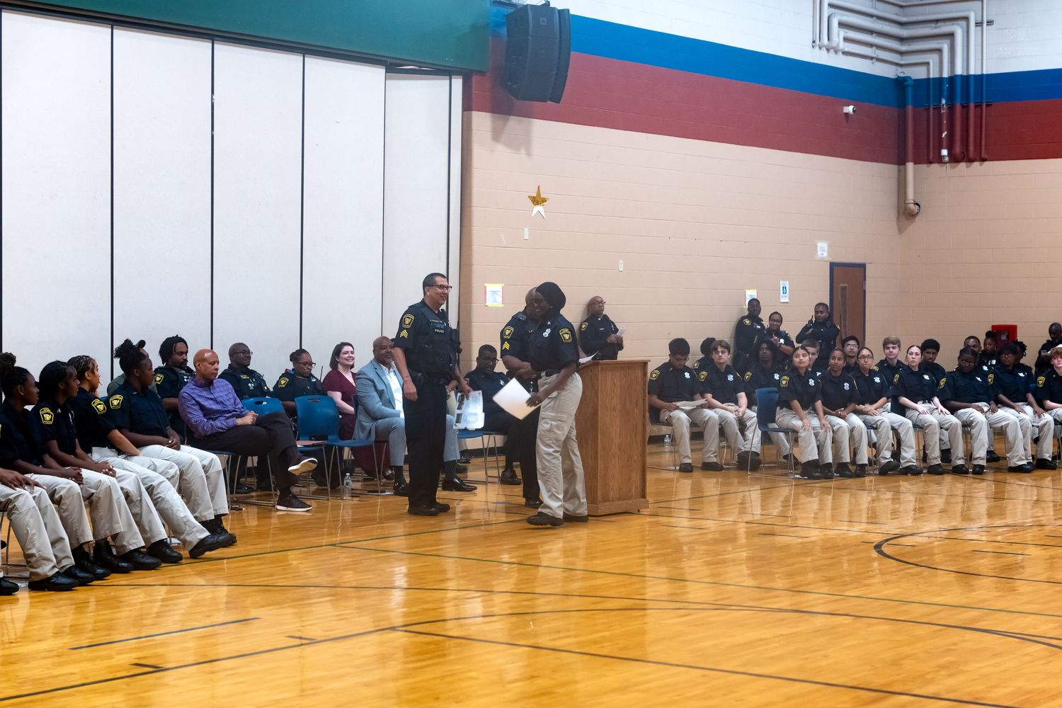 Officers and students gather in a school gymnasium for a formal ceremony, with a speaker at a podium addressing the audience.