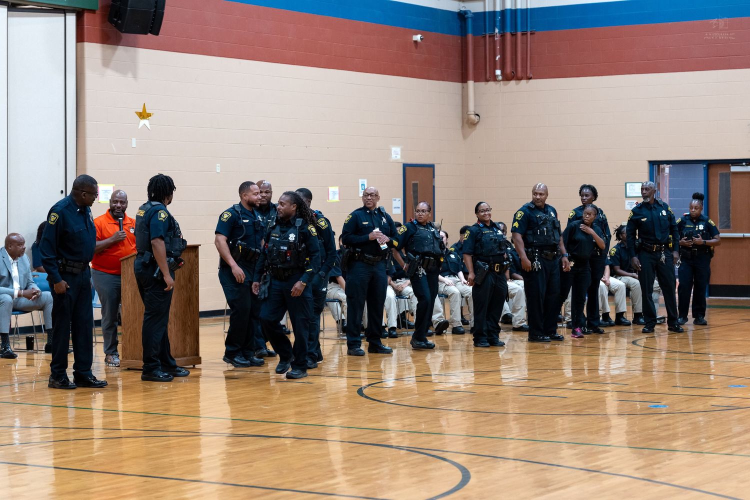 Police officers in uniform line up inside a gymnasium during a community event on a hardwood basketball court.