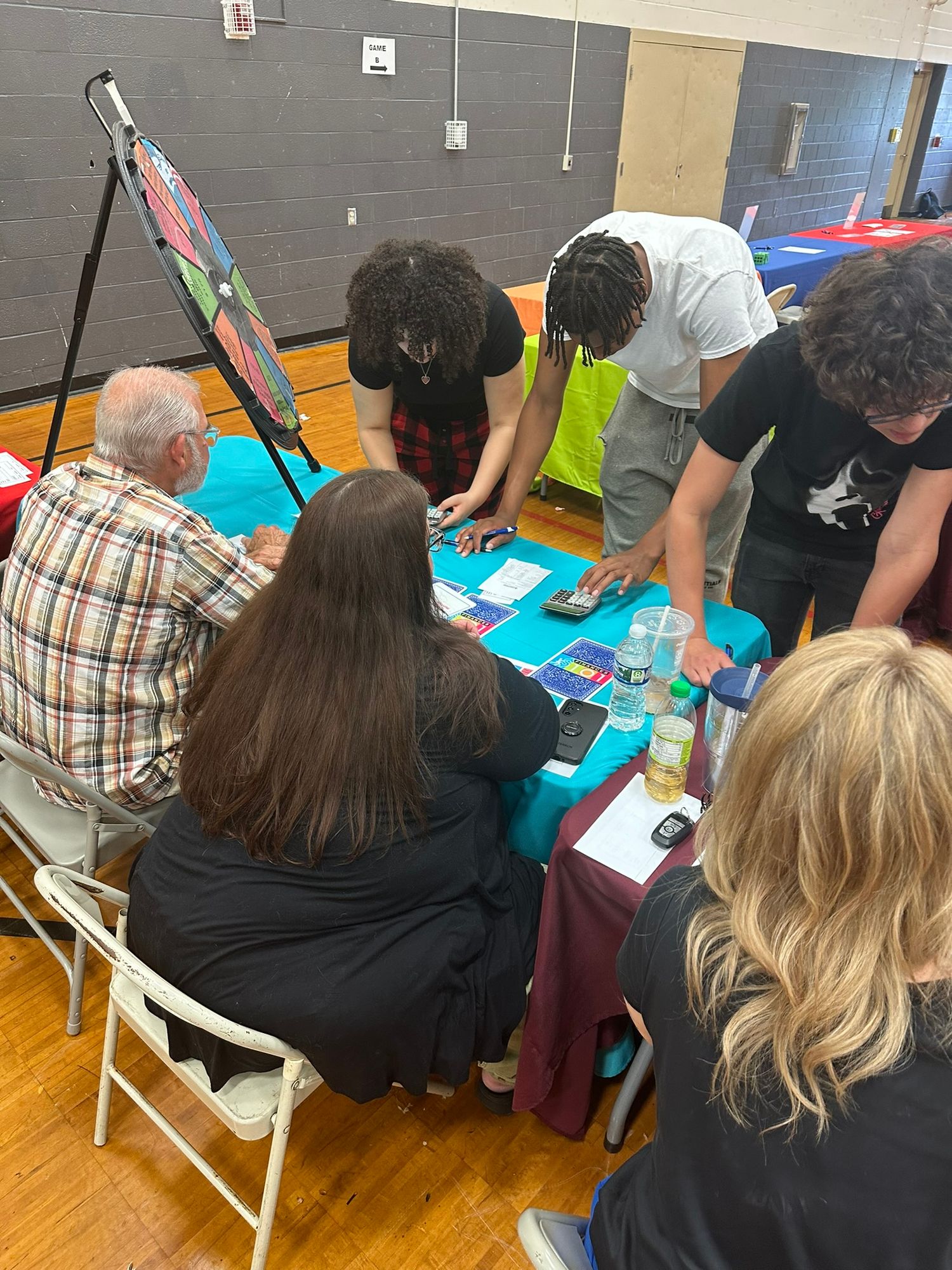 Group of people gathered around a teal-draped table reviewing materials at an indoor community expo event.