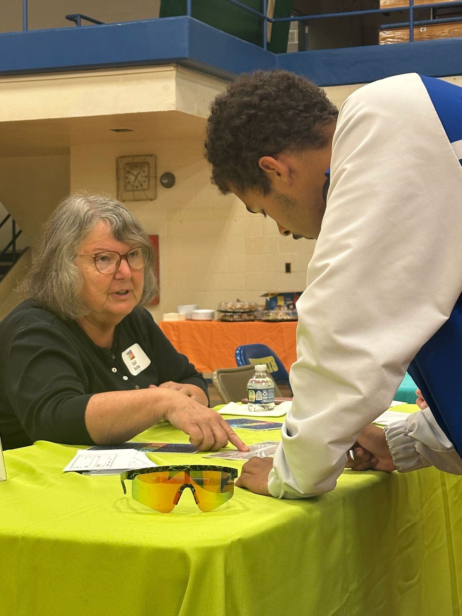 An older woman with gray hair assists a student at a lime green table during an indoor school resource fair.