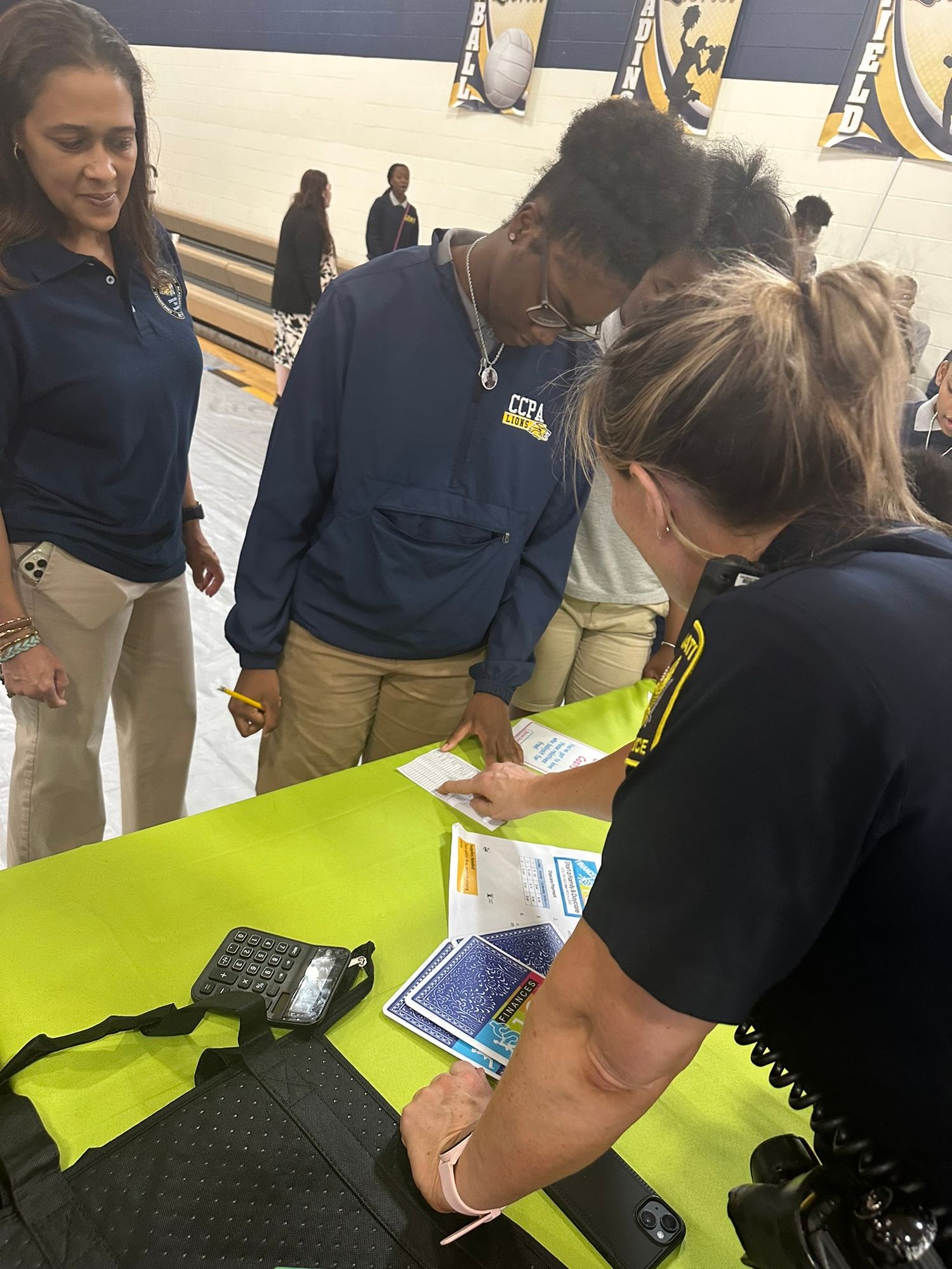 A police officer assists students at a table during a school career fair event in a gymnasium.