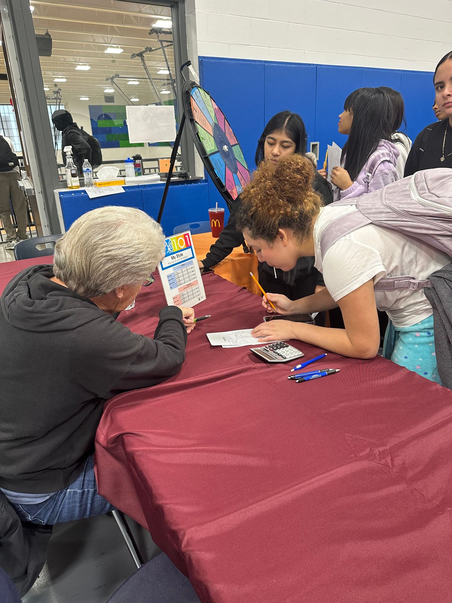 Students and an elderly person review documents at a red-draped table during a school fair event.