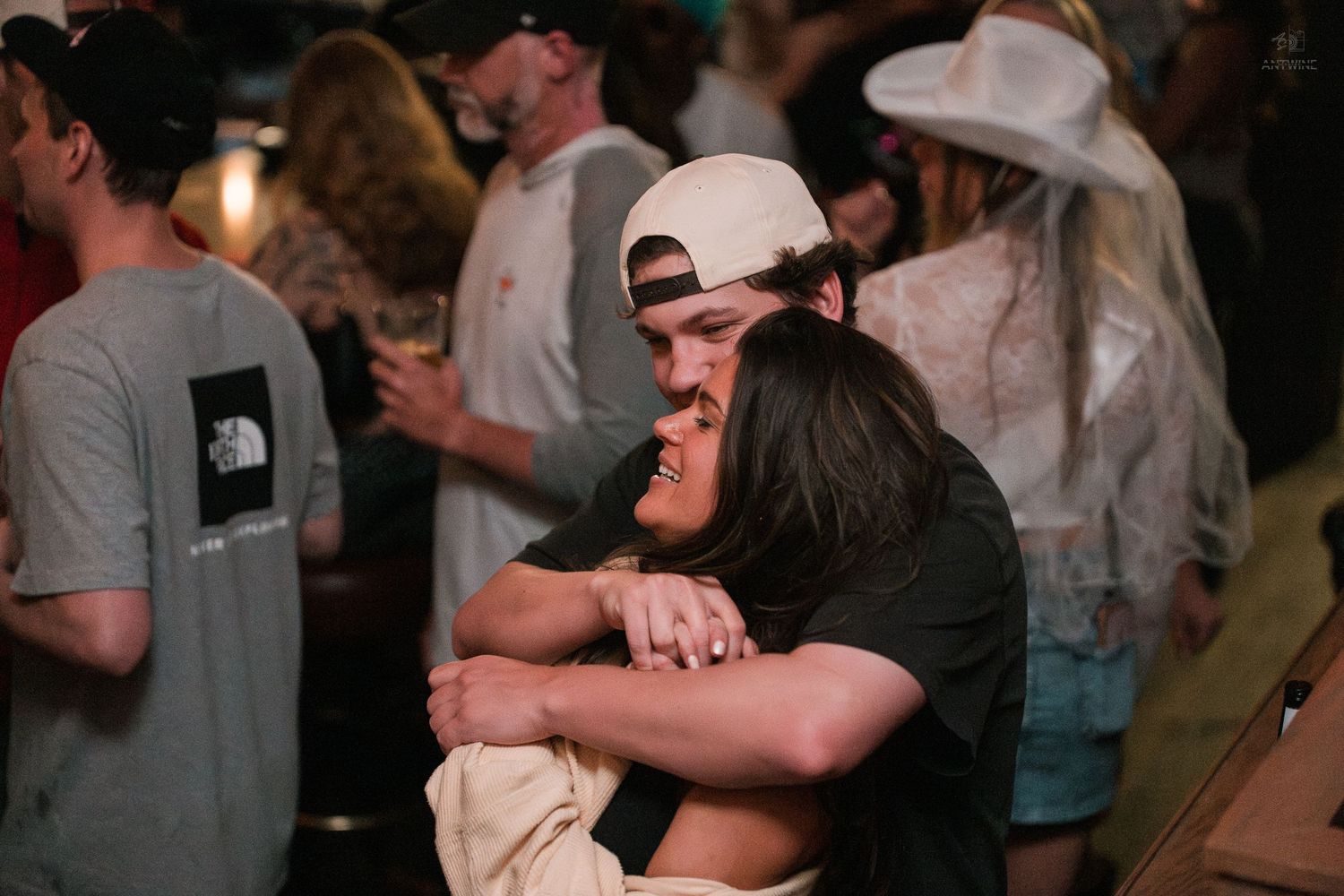 Two people sharing a warm hug at a busy bar gathering with crowd visible in background.