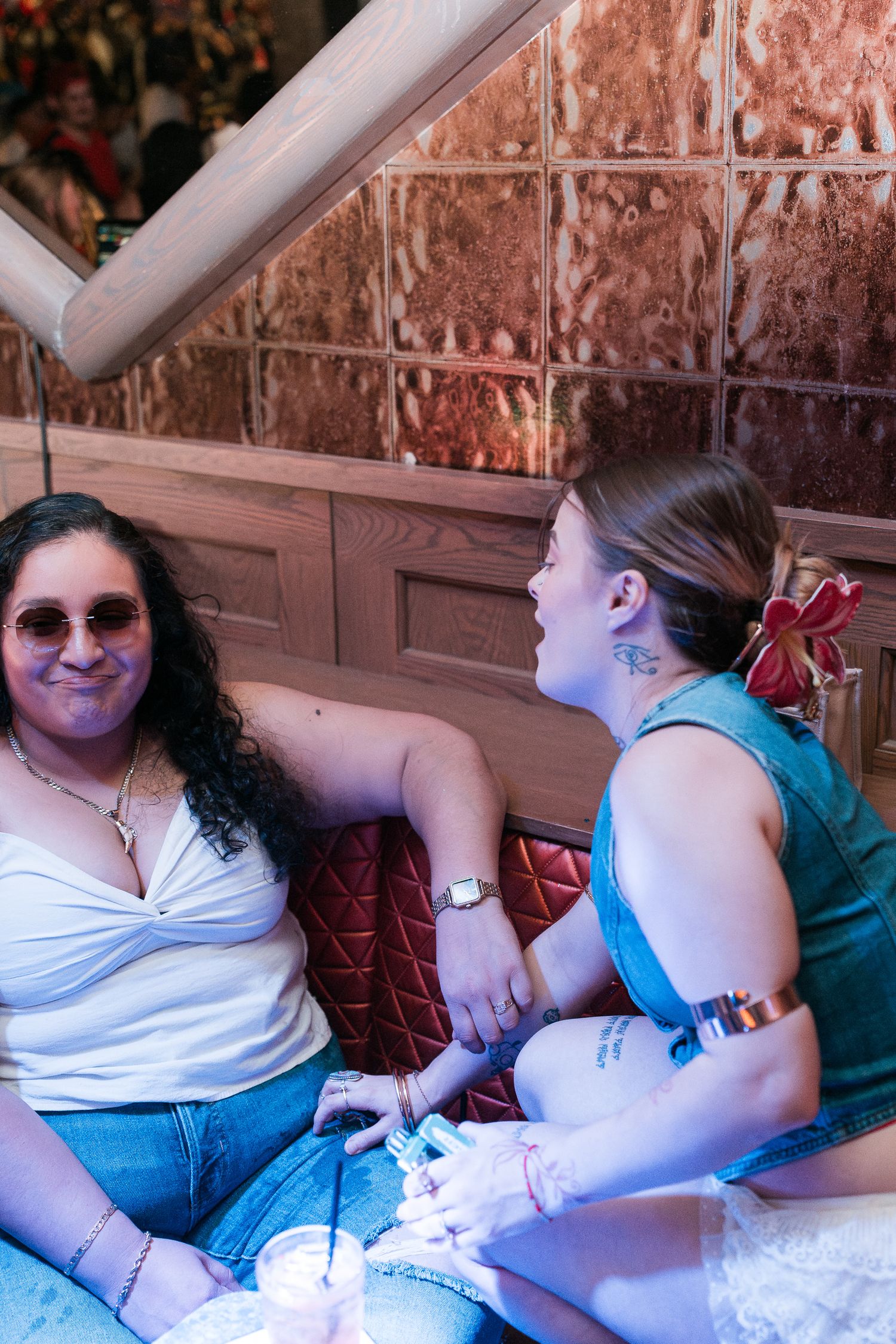 Two women chatting in a cozy bar booth with copper tile walls and red seating.