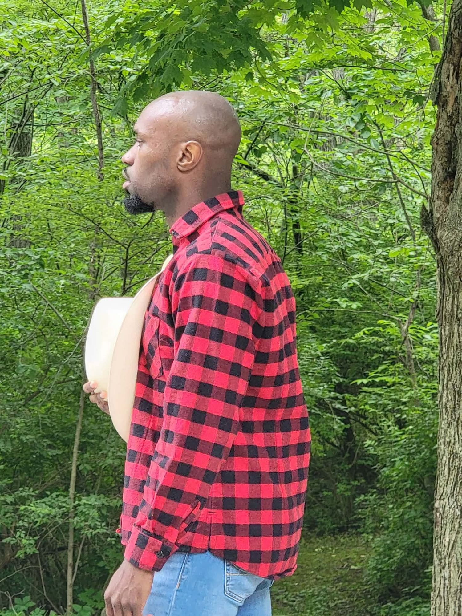 A bald Black man in a red and black plaid flannel shirt stands in profile amid lush green forest trees.