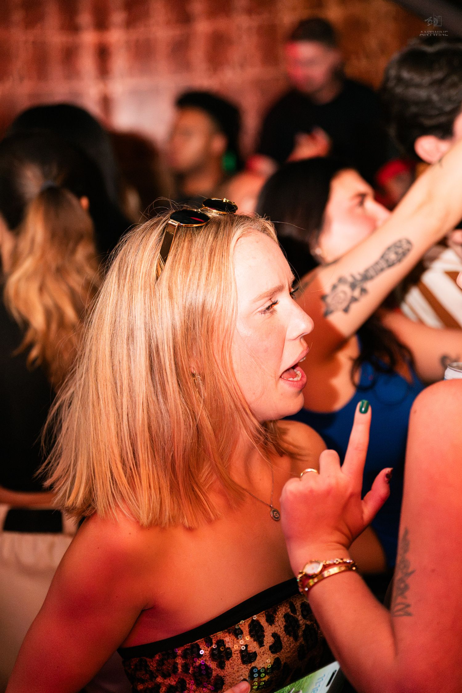 Blonde woman in leopard top gestures at lively indoor party or bar event.