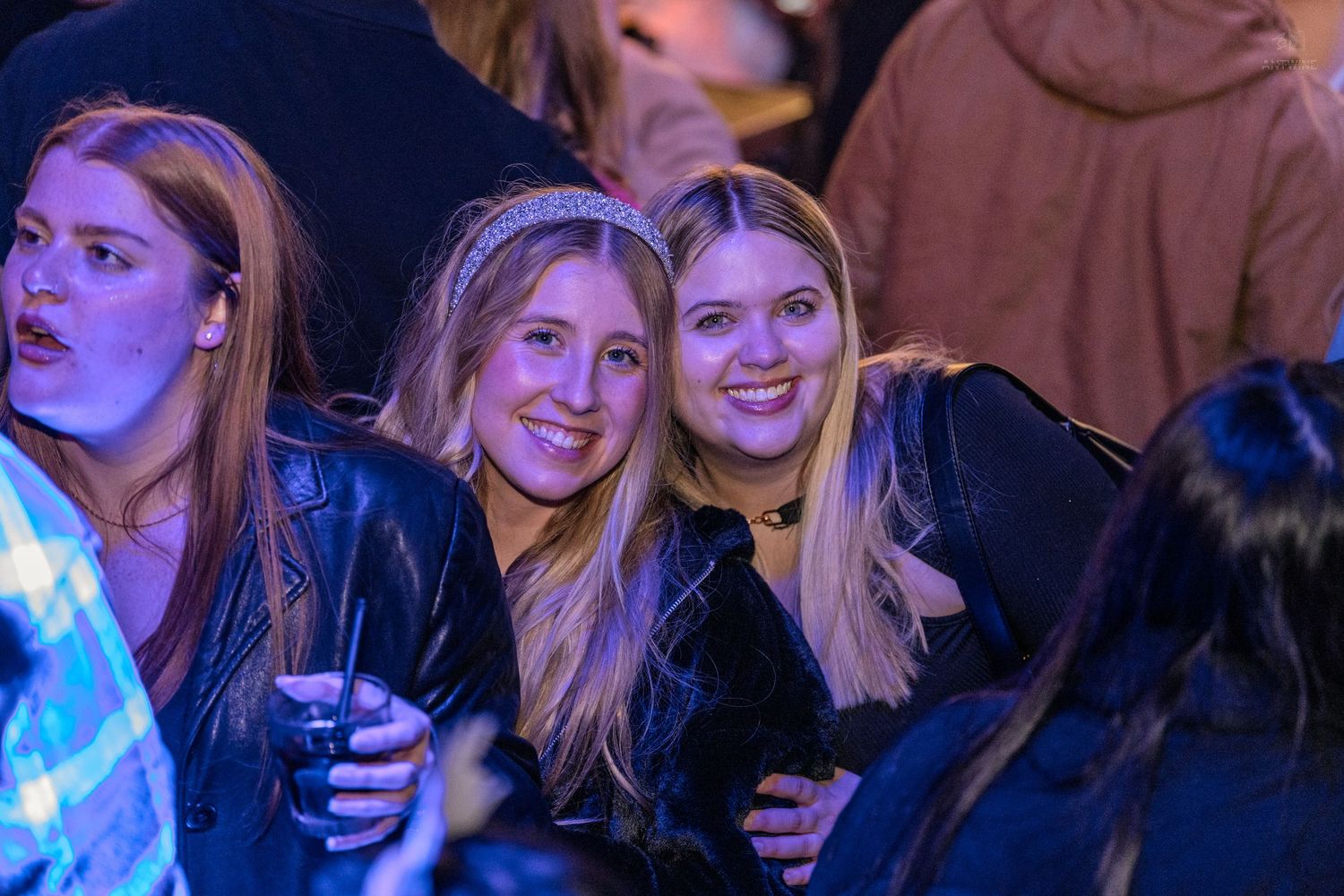 Two blonde women smile and embrace at an outdoor concert under blue stage lighting.