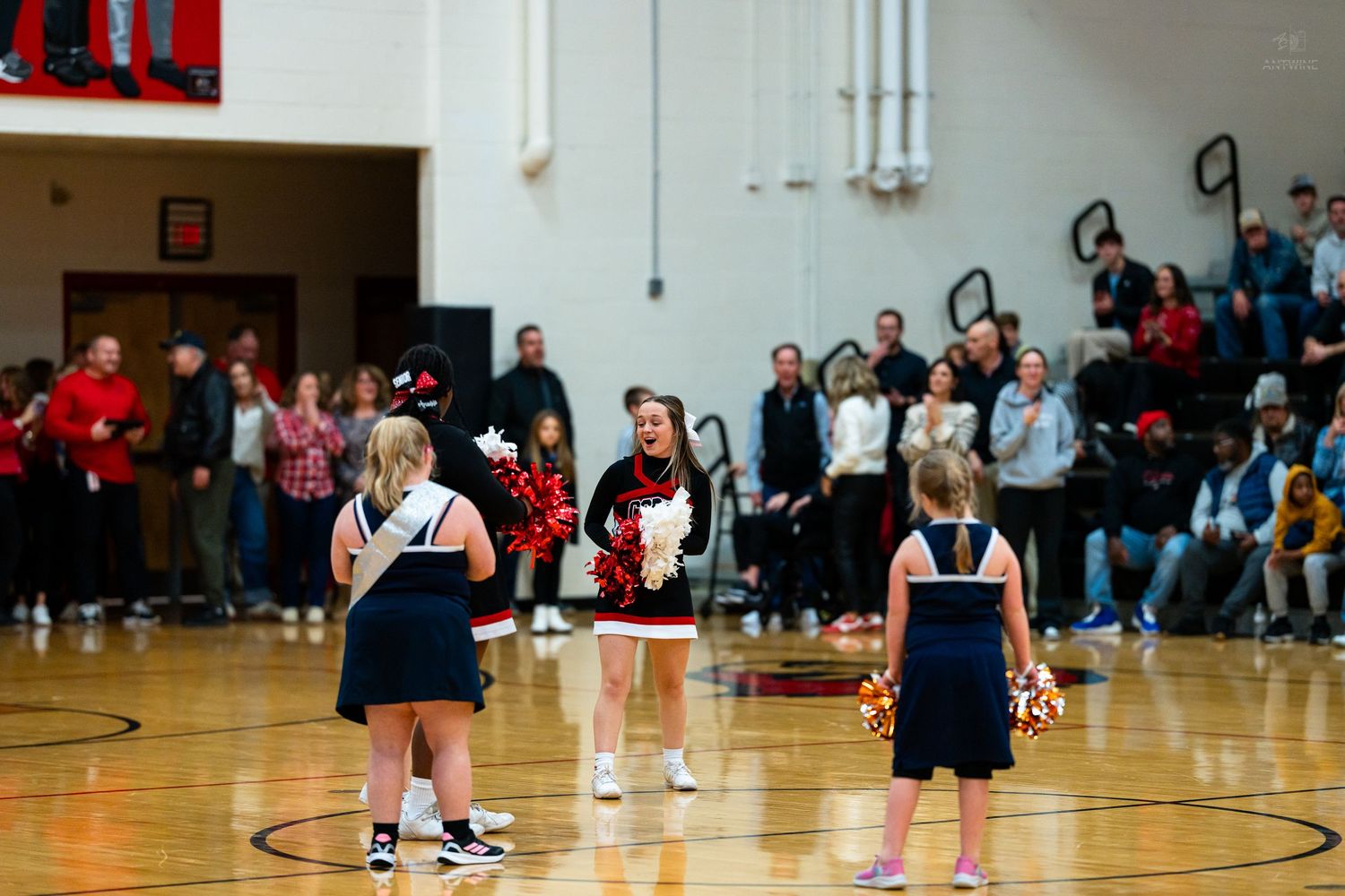 College cheerleader in red guides young cheerleaders in navy uniforms during basketball game halftime show.