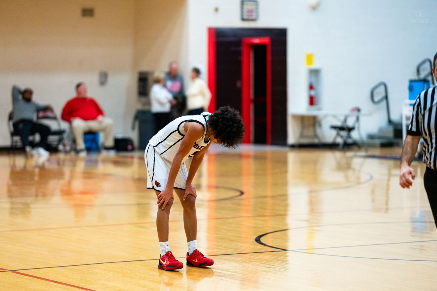 A young basketball player bends over catching breath on a hardwood court during a high school game.
