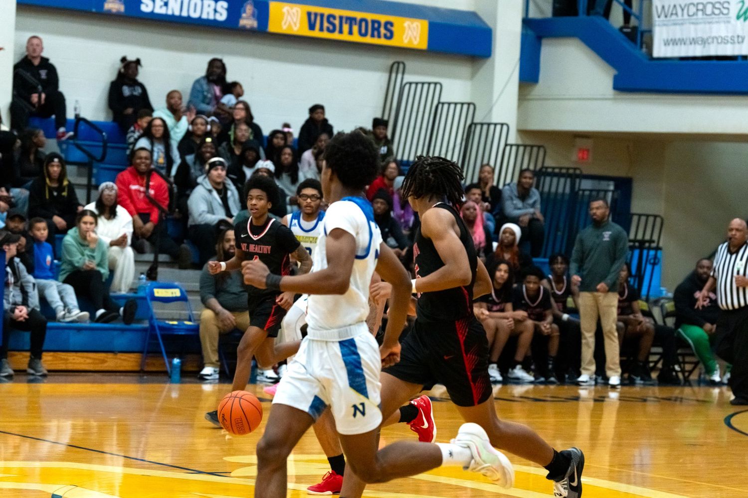 High school basketball player dribbles past defenders near the sideline in a crowded gymnasium.