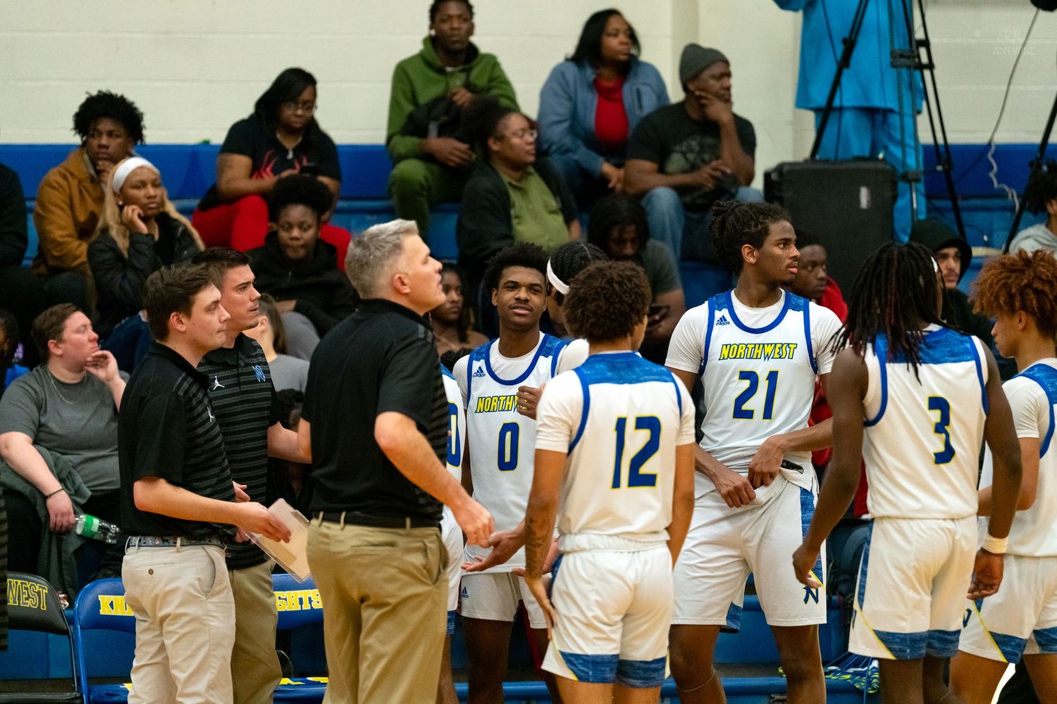 High school basketball coach talks to his team during a timeout in a blue and white gymnasium.