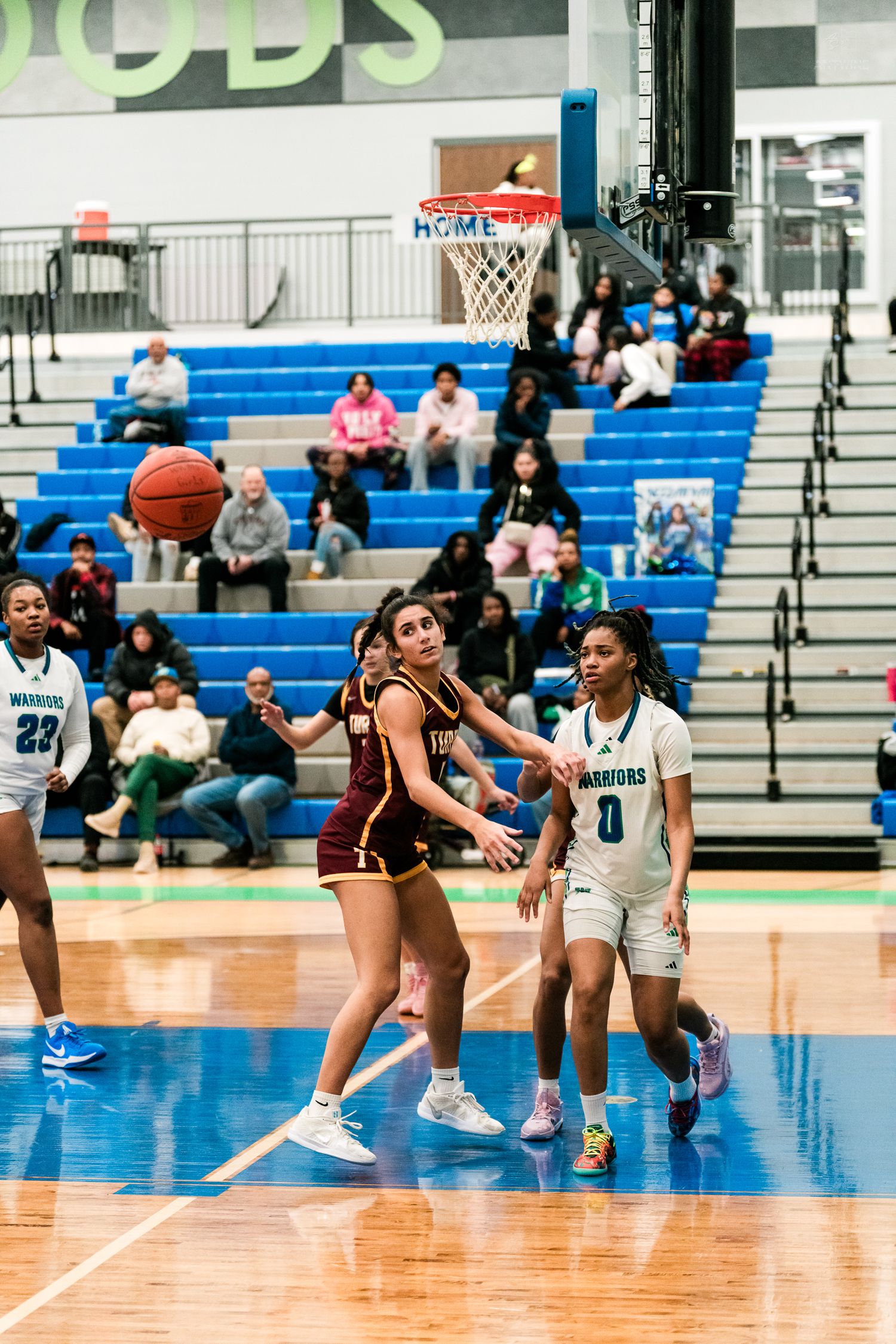 Female basketball player in maroon uniform passes the ball while teal-uniformed Warriors defenders close in near the basket.