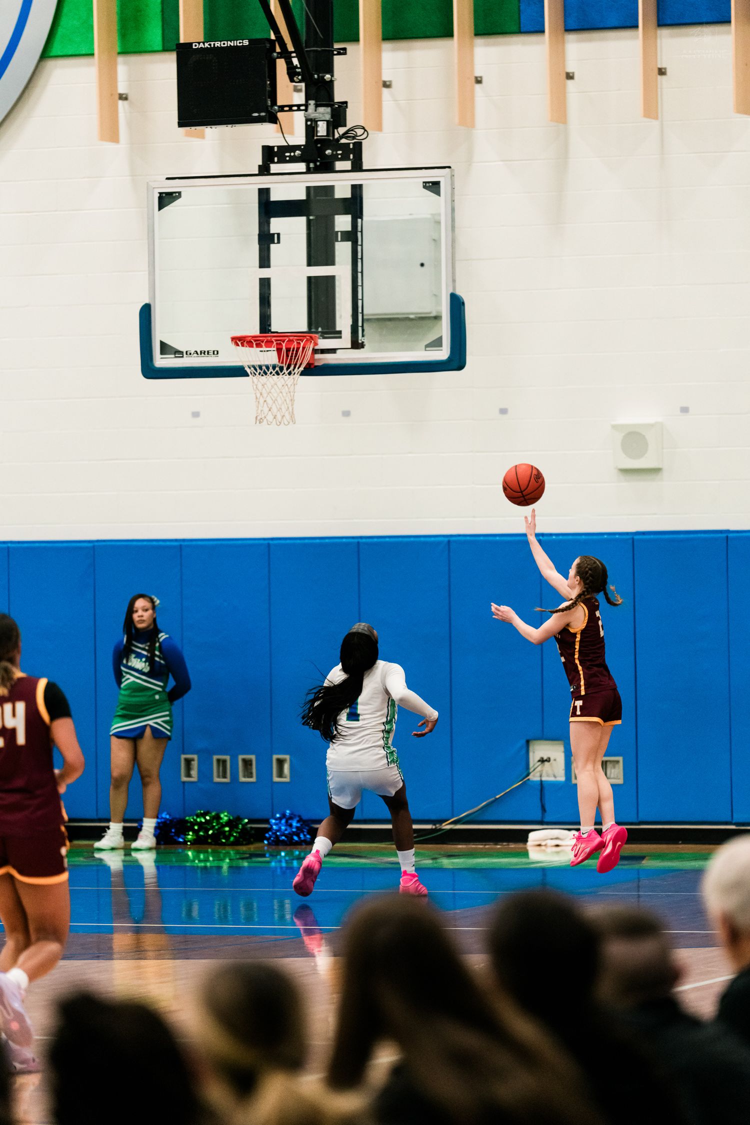 Maroon-uniformed player shoots a jump shot near the basket while a white-uniformed defender contests during a girls basketball game.