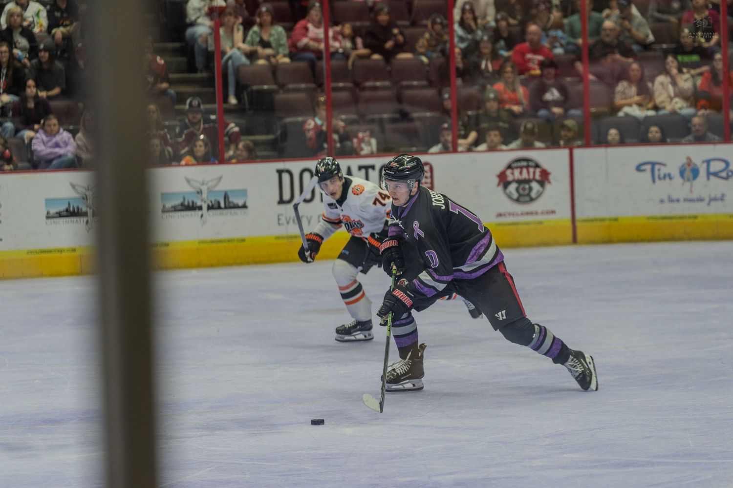 Hockey player in dark uniform skates with puck while orange-jersied opponent pursues during indoor ice hockey game.