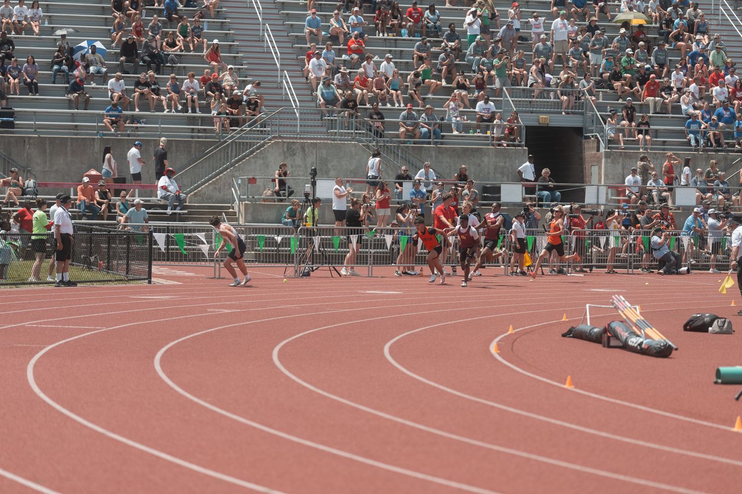 Athletes compete in a track and field relay race at a packed outdoor stadium on a sunny day.