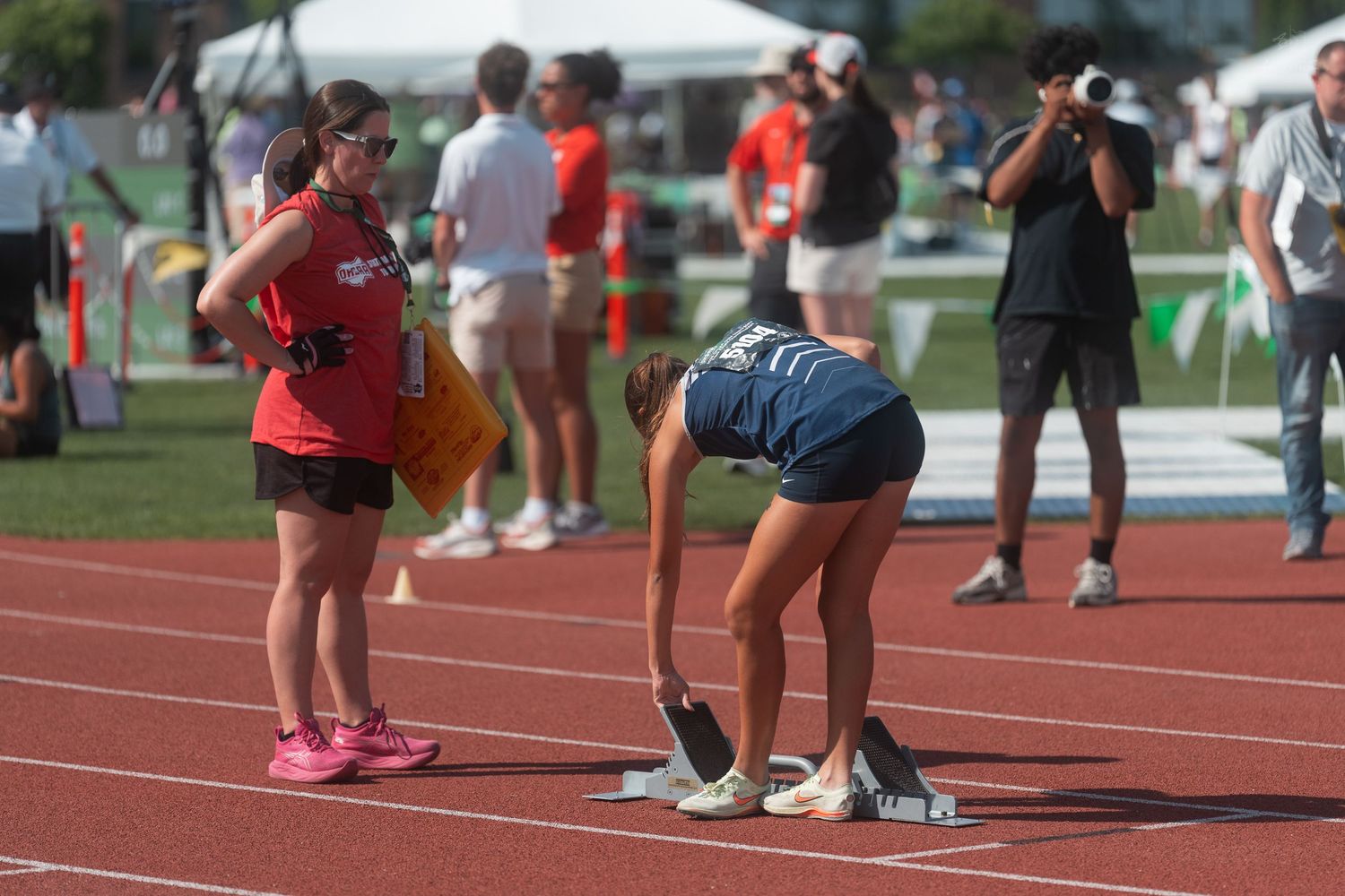 Female track athlete sets starting blocks on a red track while a coach in red observes nearby.