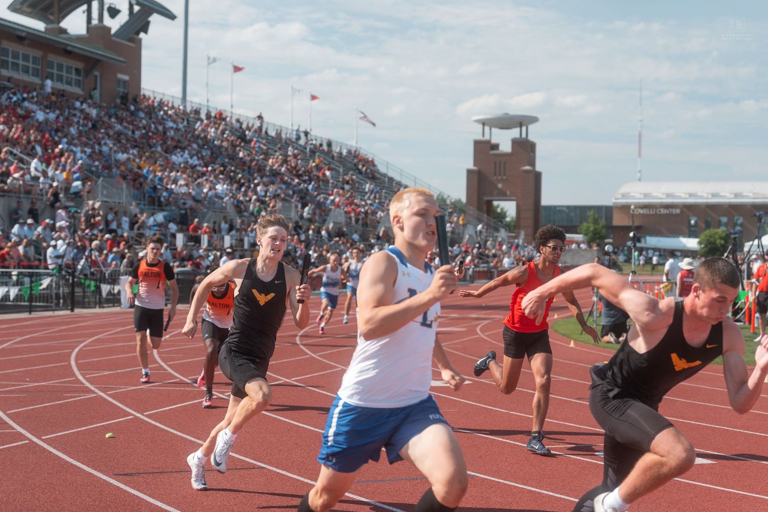 Runners compete in a relay race at a large outdoor track meet with packed stadium bleachers in the background.