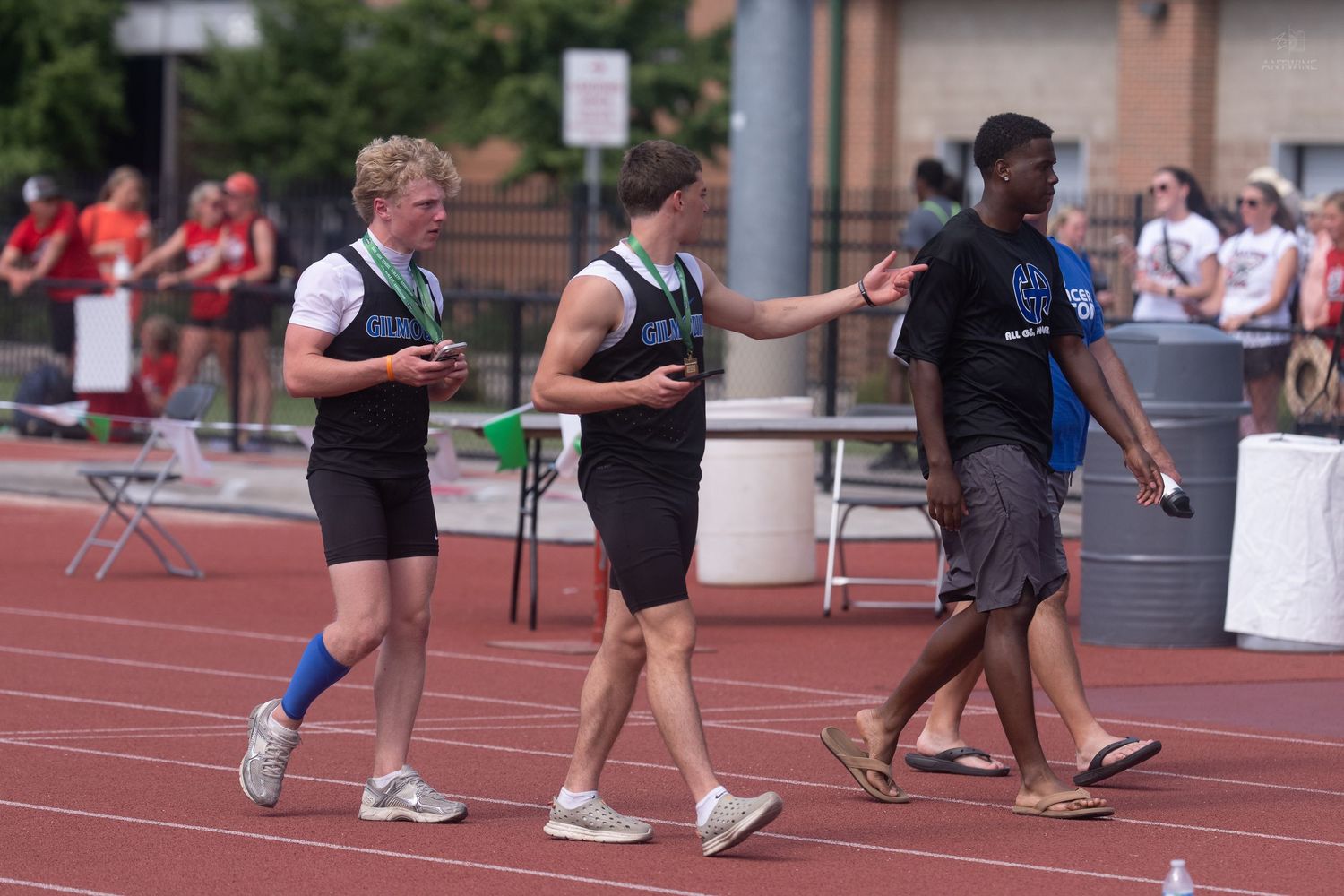 Three male track athletes celebrate on a red track after completing a relay race at an outdoor meet.