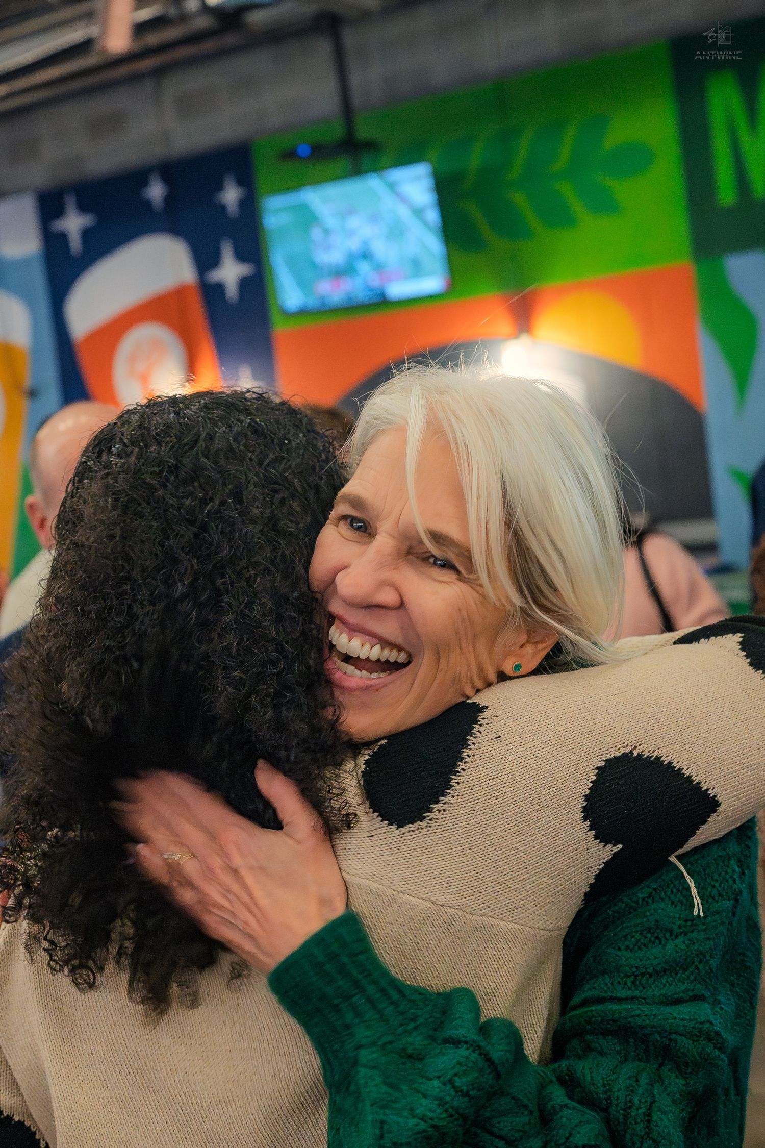 Two women share a warm embrace and joyful smile at a colorful indoor event.