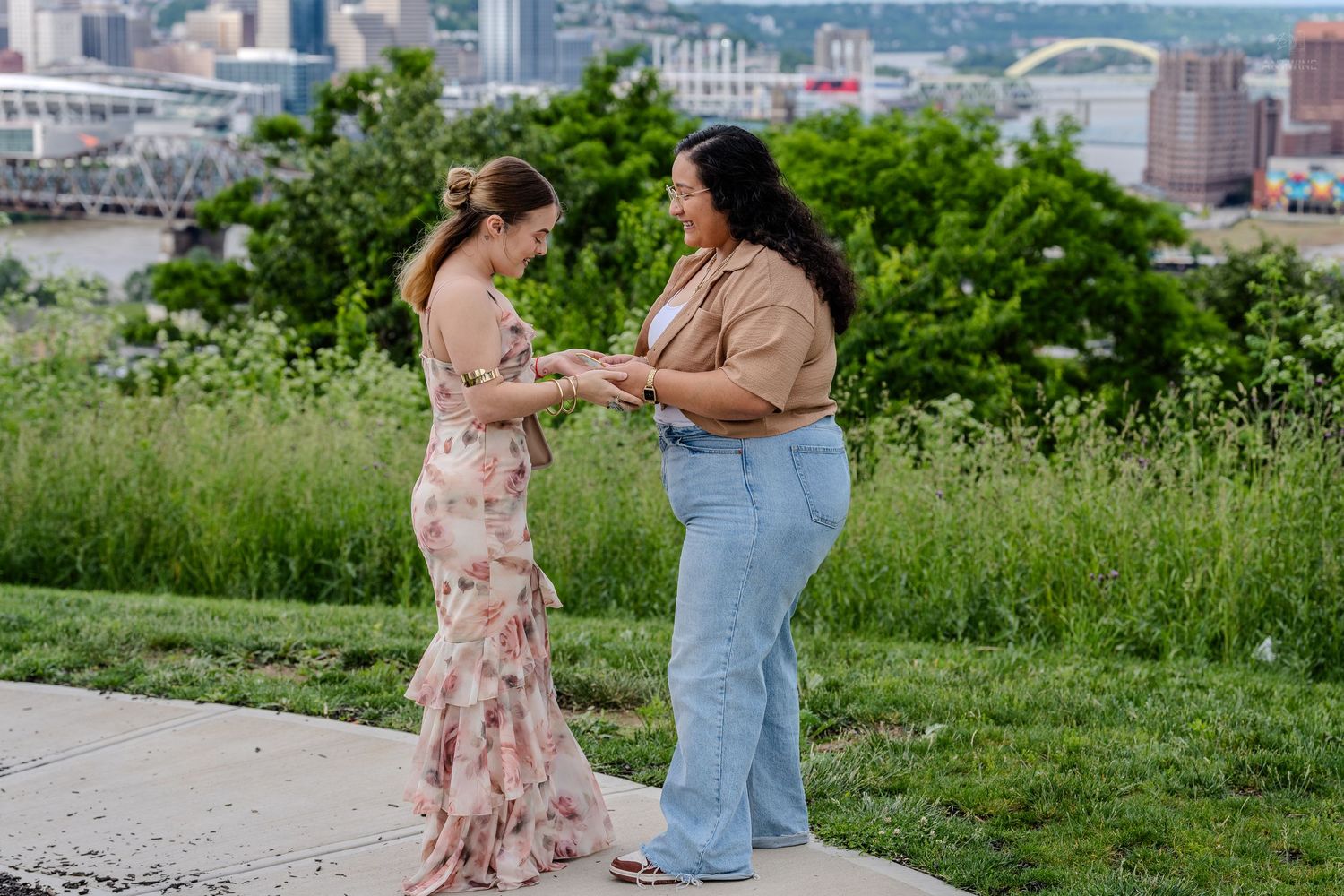 Two women holding hands during marriage proposal on scenic overlook with Cincinnati skyline behind.