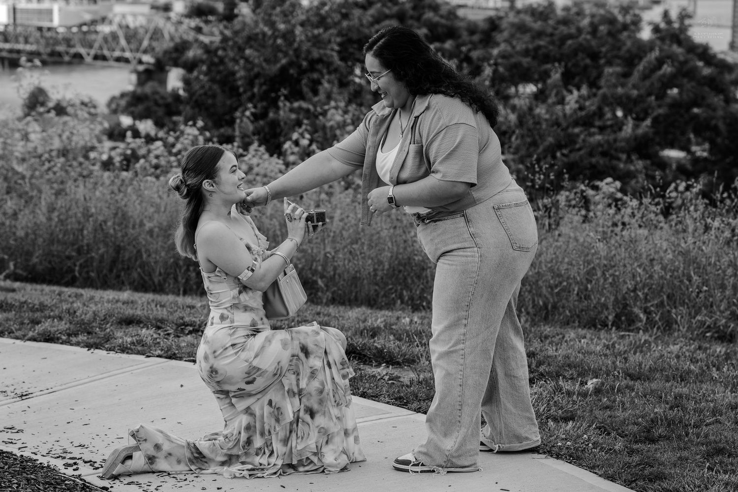 Woman on one knee proposing with ring to partner on outdoor pathway.