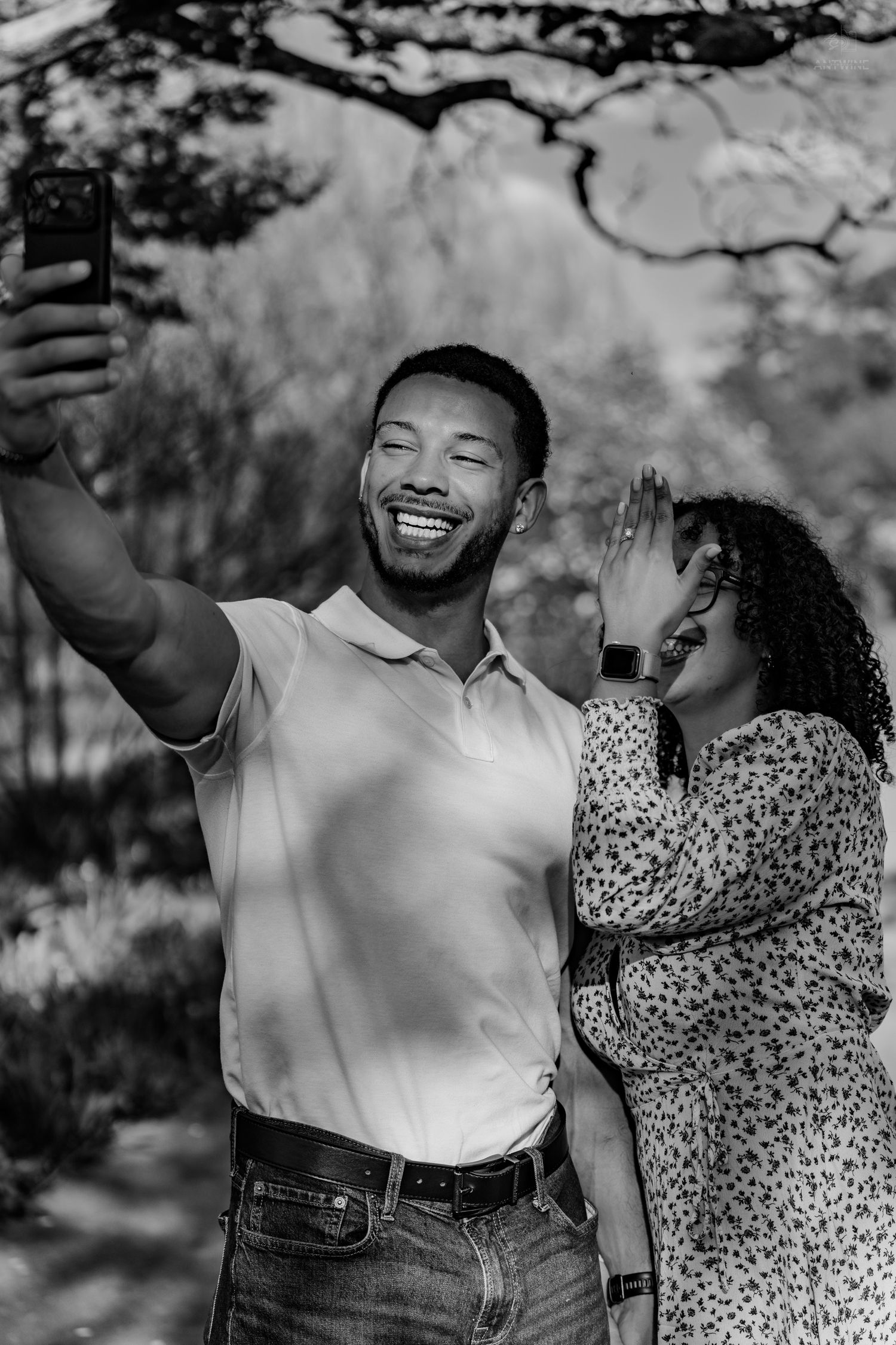 Black and white photo of happy couple taking a selfie outdoors, woman waving engagement ring hand at camera.